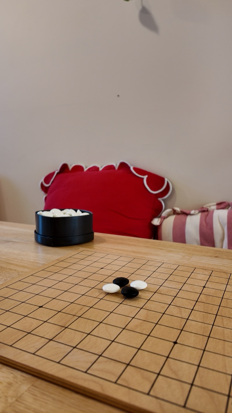 un goban en bois sur une table du café, quatre pierres posées, un bol de pierres blanches et un coussin rouge derrière