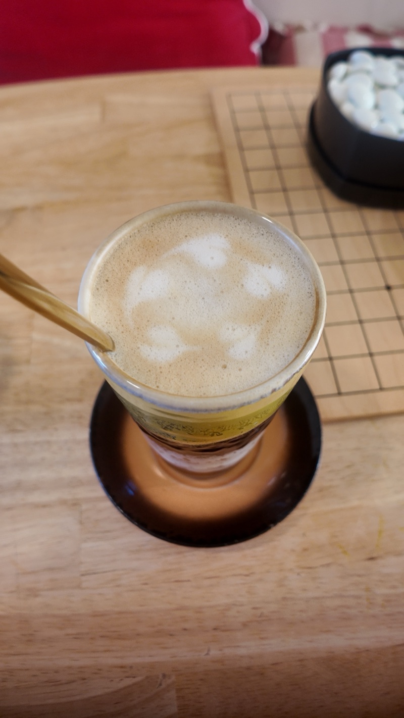 un café crème à la cuillère dorée posé à côté du goban, un bol de pierres blanches en arrière-plan